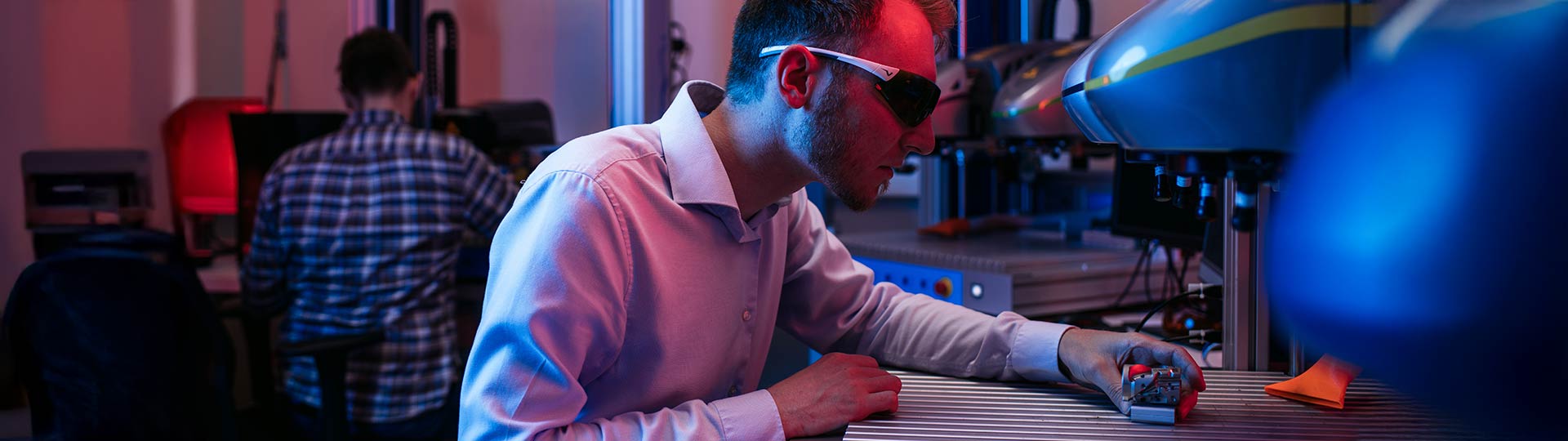 Bediener mit Laserschutzbrille platziert Bauteil unter Beschriftungslaser mit Vision-System in Laborumgebung mit mehreren Lasersystemen